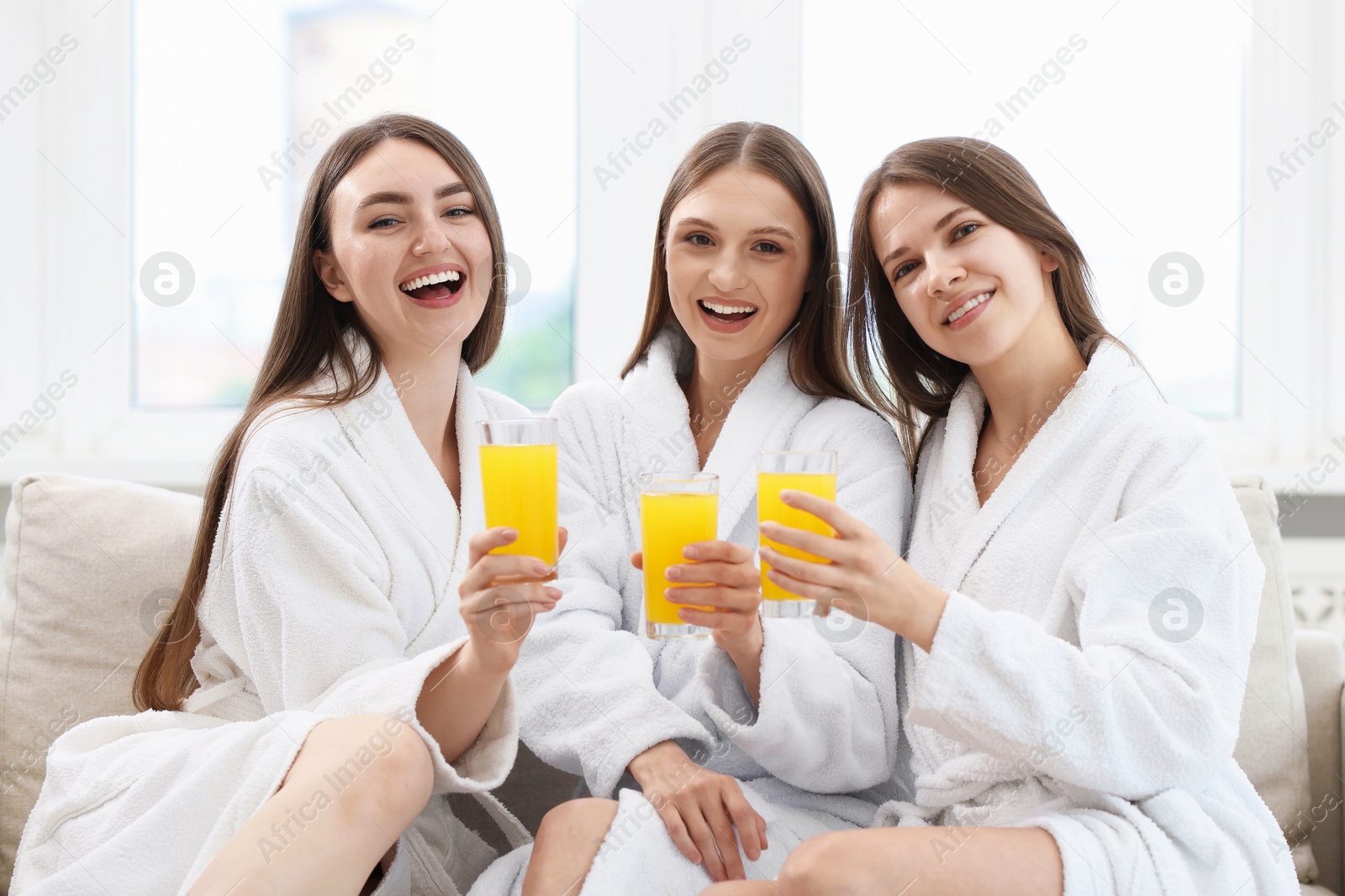 Photo of Happy women in bathrobes with glasses of orange juice on sofa in spa salon