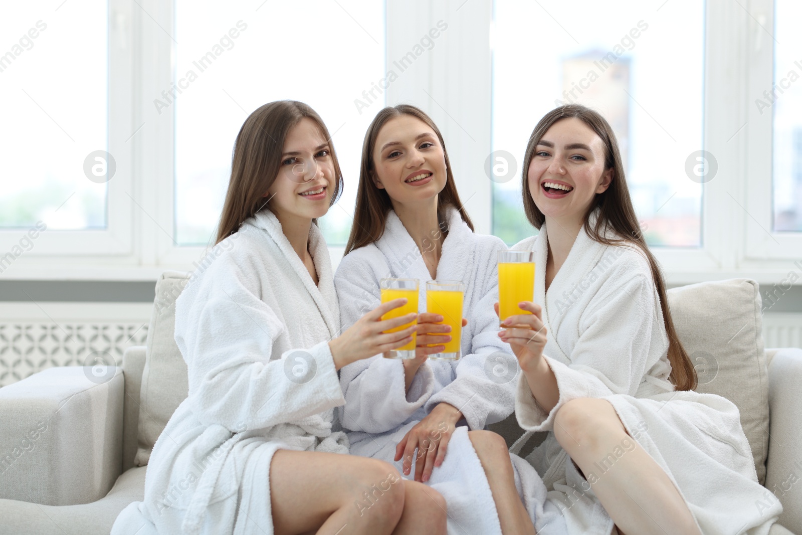 Photo of Happy women in bathrobes with glasses of orange juice on sofa in spa salon
