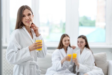 Photo of Smiling woman in bathrobe with glass of orange juice and her friends in spa salon, selective focus
