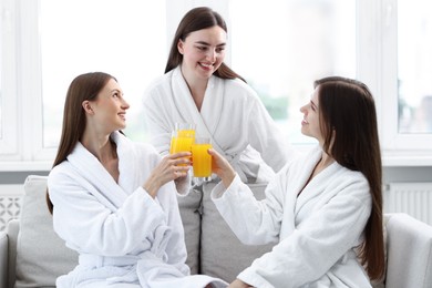 Photo of Happy women in bathrobes clinking glasses of orange juice on sofa in spa salon