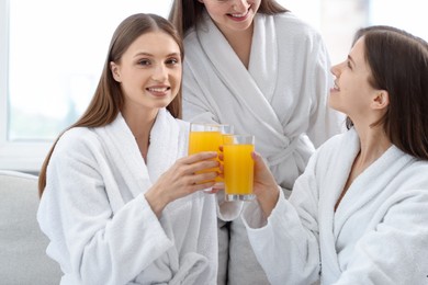 Photo of Happy women in bathrobes clinking glasses of orange juice in spa salon, closeup