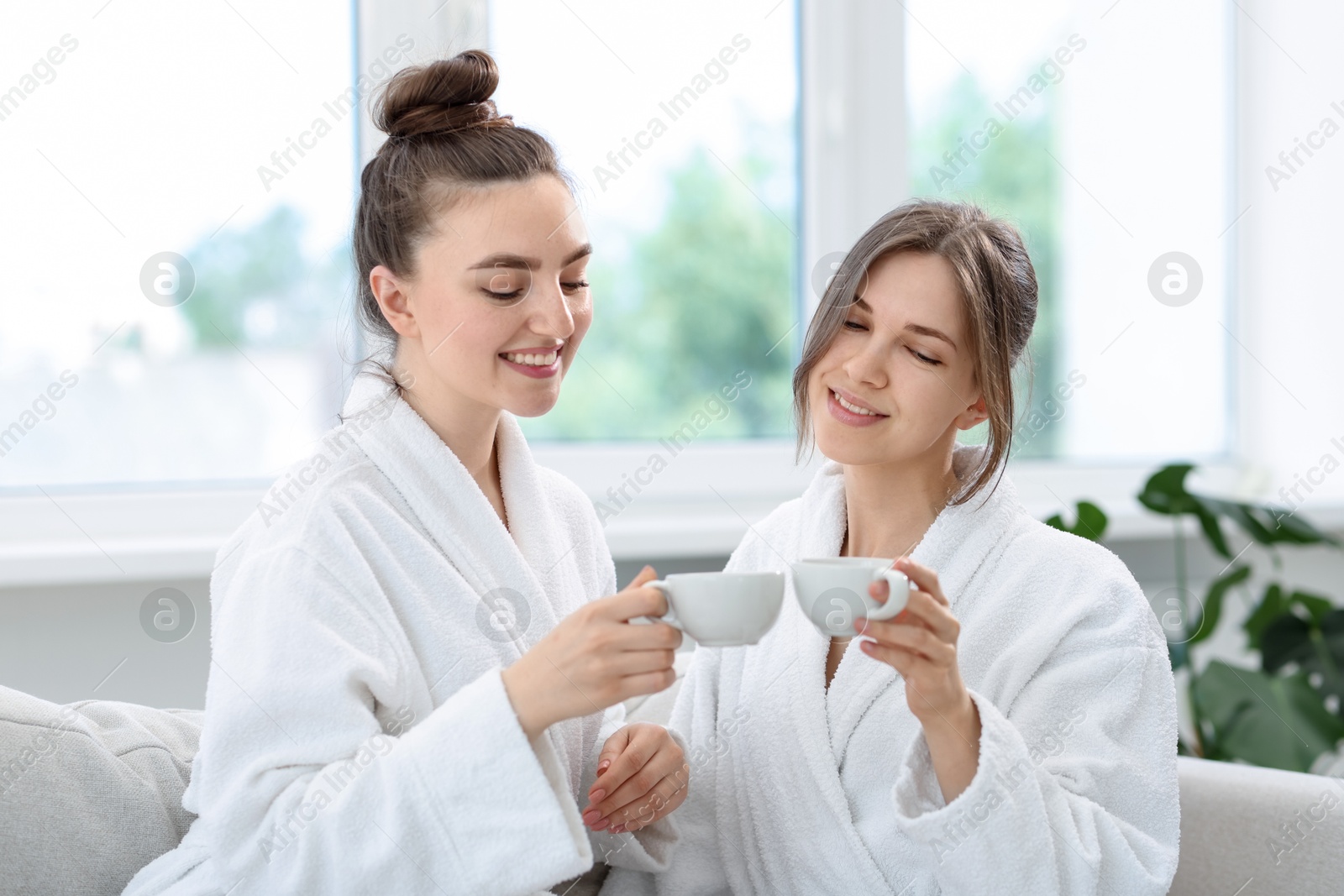 Photo of Happy women in bathrobes with cups of healthy drink after spa procedures indoors
