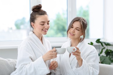 Photo of Happy women in bathrobes with cups of healthy drink after spa procedures indoors