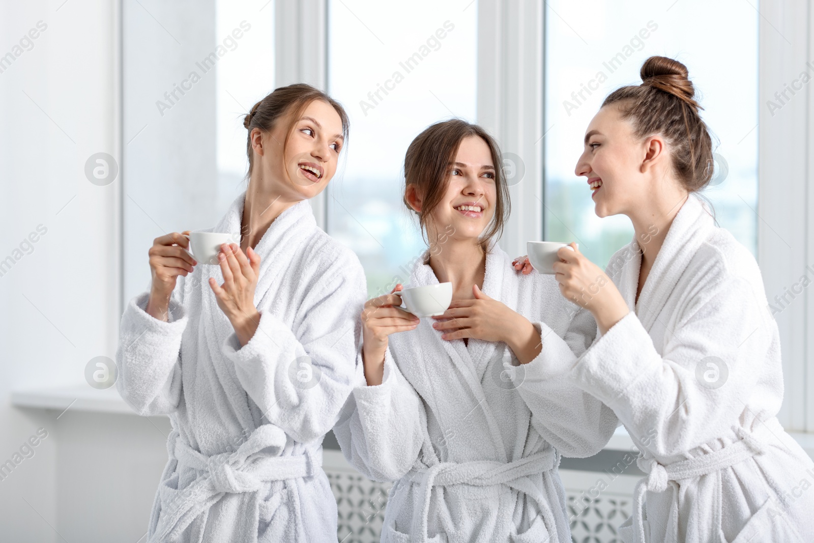 Photo of Happy women in bathrobes with cups of healthy drink after spa procedures indoors