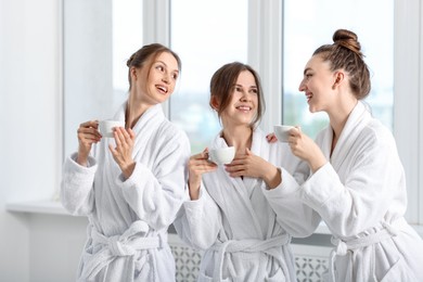 Photo of Happy women in bathrobes with cups of healthy drink after spa procedures indoors