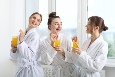 Photo of Happy women in bathrobes with glasses of orange juice after spa procedures indoors