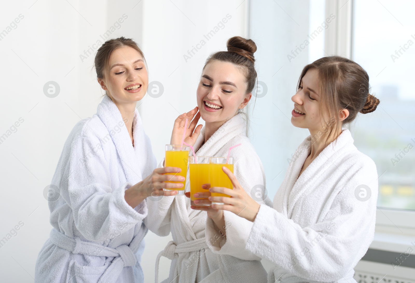 Photo of Happy women in bathrobes clinking glasses of orange juice after spa procedures indoors