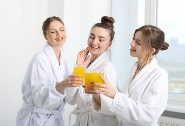 Photo of Happy women in bathrobes clinking glasses of orange juice after spa procedures indoors