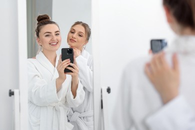 Photo of Happy women in bathrobes taking selfie near mirror after spa procedures indoors