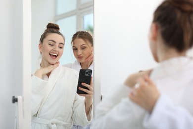 Photo of Happy women in bathrobes taking selfie near mirror after spa procedures indoors