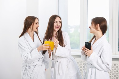 Photo of Happy women in bathrobes with glasses of orange juice and smartphone in spa salon