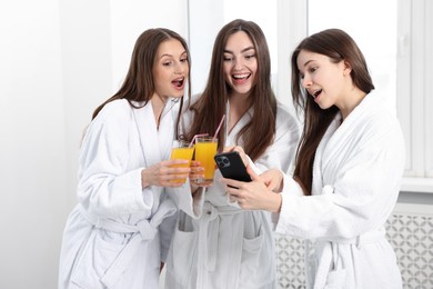 Photo of Happy women in bathrobes with glasses of orange juice looking at smartphone in spa salon
