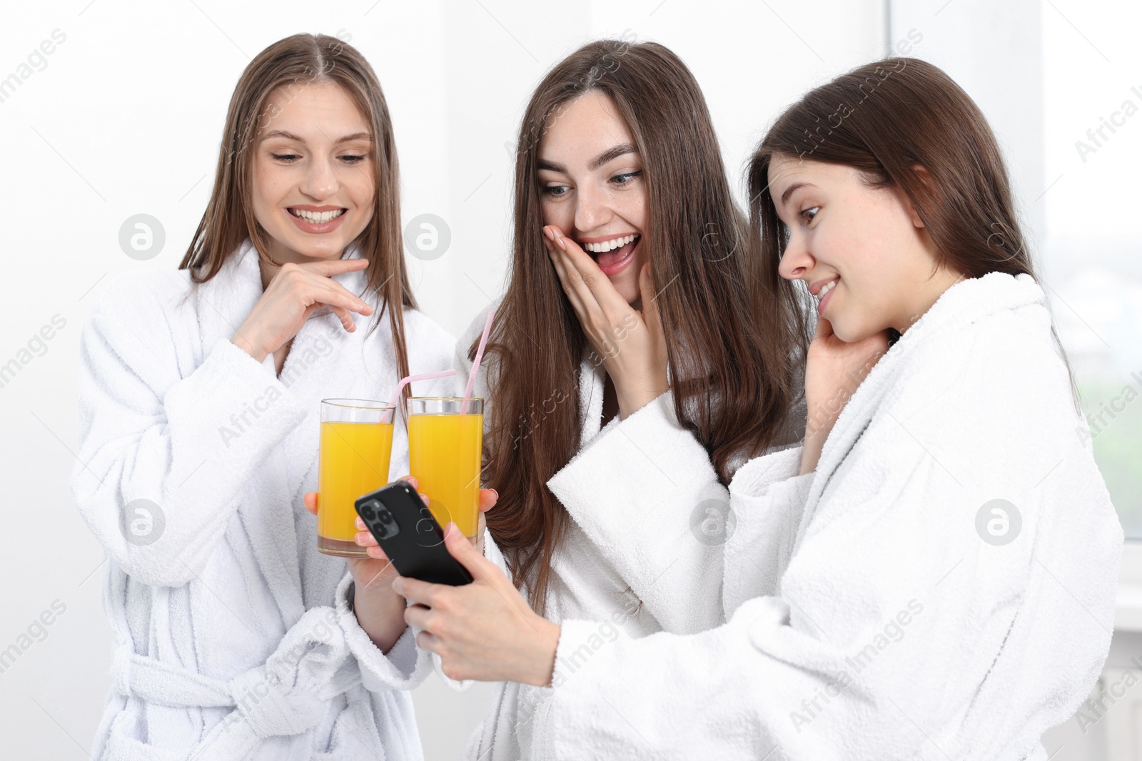 Photo of Happy women in bathrobes with glasses of orange juice looking at smartphone in spa salon