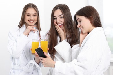 Photo of Happy women in bathrobes with glasses of orange juice looking at smartphone in spa salon