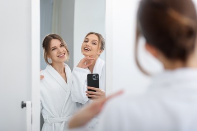 Photo of Happy women in bathrobes taking selfie near mirror after spa procedures indoors