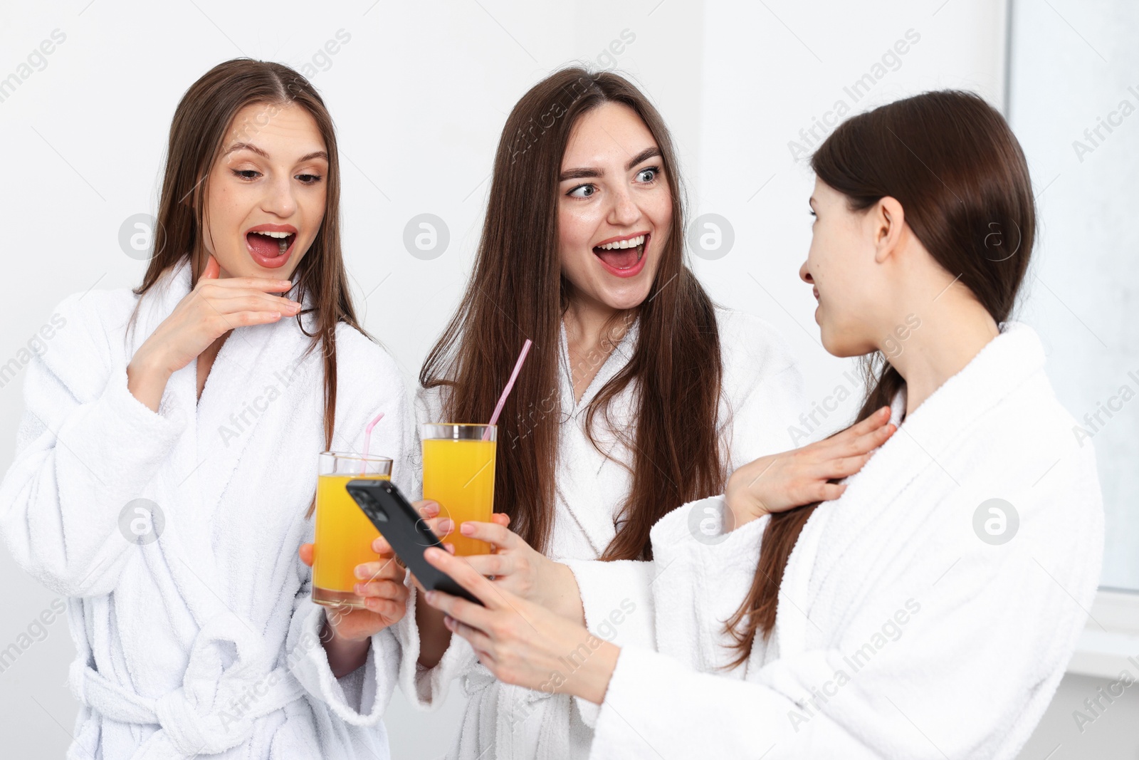Photo of Happy women in bathrobes with glasses of orange juice and smartphone in spa salon