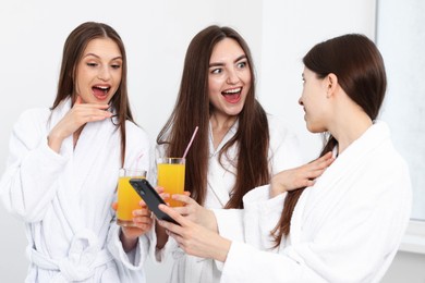 Photo of Happy women in bathrobes with glasses of orange juice and smartphone in spa salon