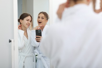 Photo of Happy women in bathrobes taking selfie near mirror after spa procedures indoors