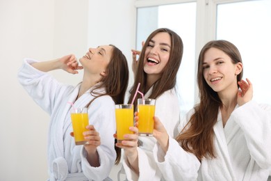 Photo of Happy women in bathrobes with glasses of orange juice after spa procedures indoors
