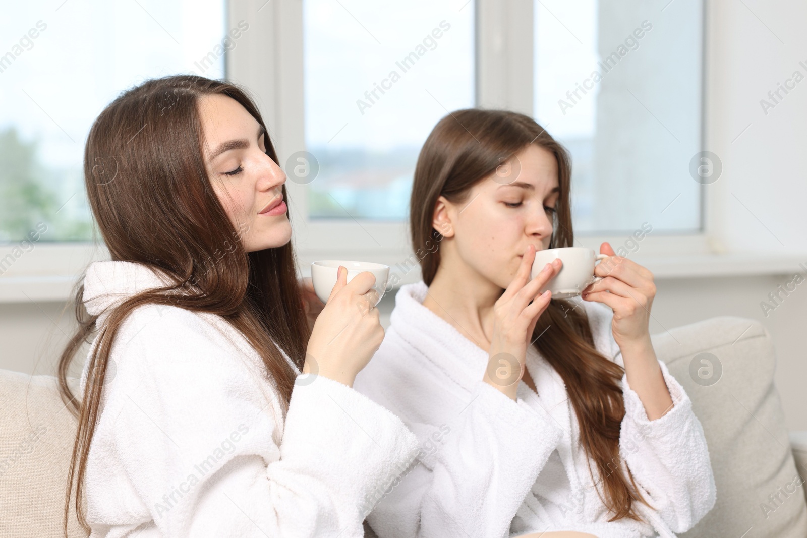 Photo of Beautiful women in bathrobes with cups of healthy drink after spa procedures on sofa indoors