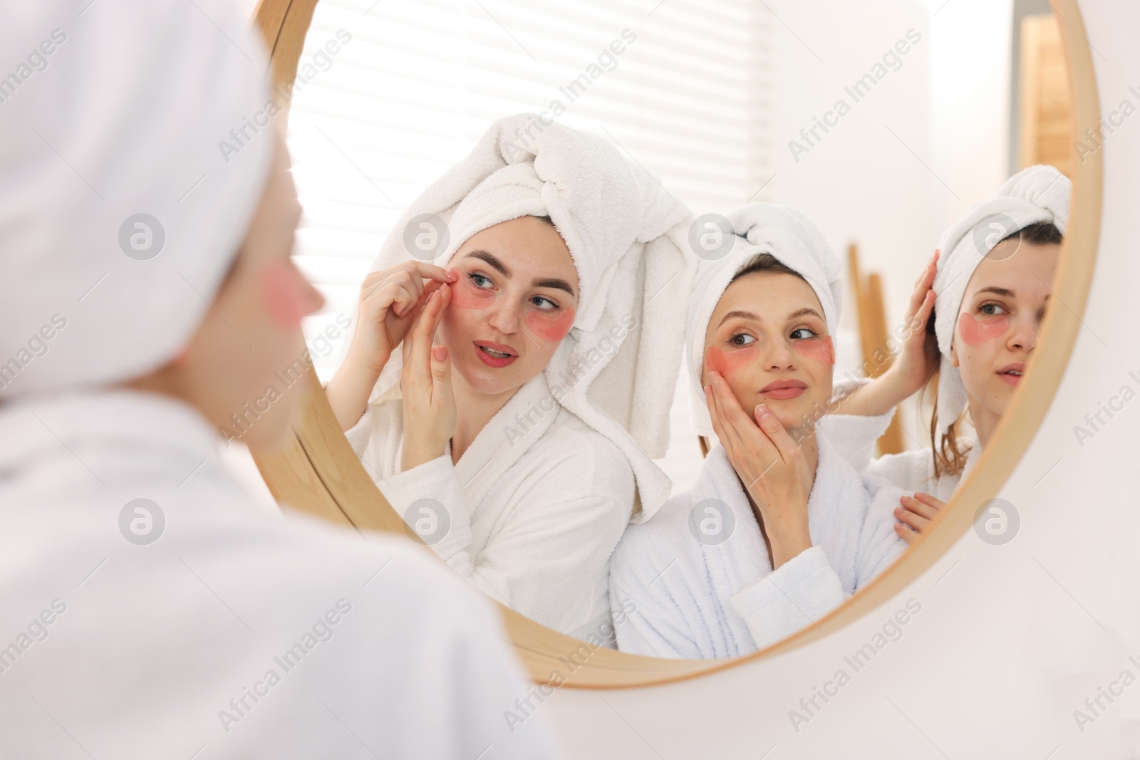 Photo of Happy women in bathrobes applying eye patches after spa procedures near mirror indoors