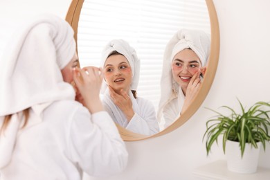 Photo of Happy women in bathrobes applying eye patches after spa procedures near mirror indoors