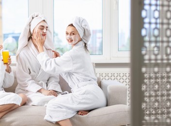 Photo of Happy women in bathrobes applying facial masks after spa procedures on sofa indoors