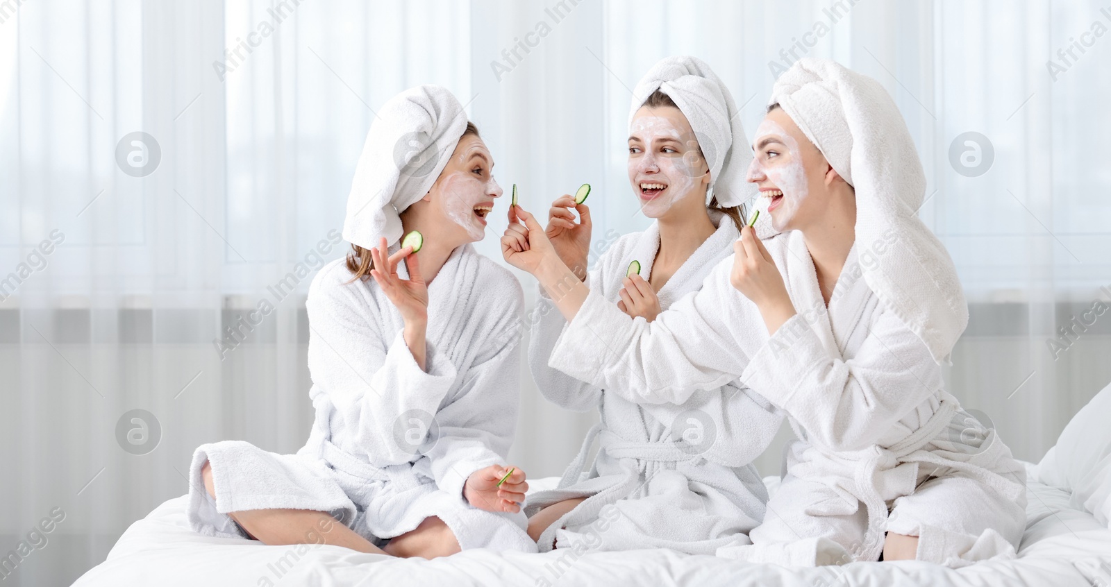 Photo of Happy women with facial masks and cucumber slices in bathrobes after spa procedures on bed indoors