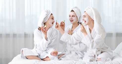 Photo of Happy women with facial masks and cucumber slices in bathrobes after spa procedures on bed indoors