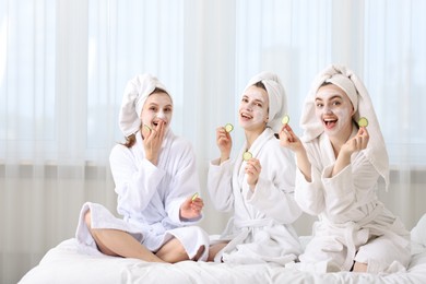 Photo of Happy women with facial masks and cucumber slices in bathrobes after spa procedures on bed indoors