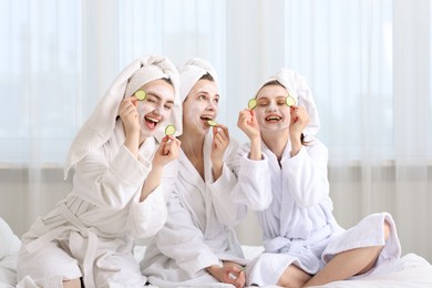 Photo of Happy women with facial masks and cucumber slices in bathrobes after spa procedures on bed indoors