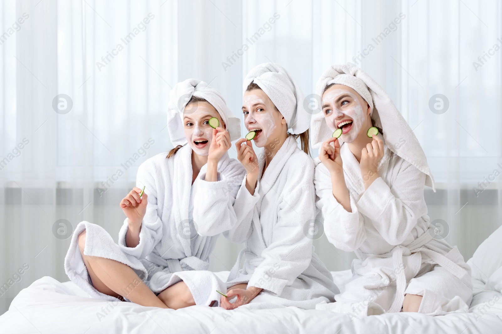Photo of Happy women with facial masks and cucumber slices in bathrobes after spa procedures on bed indoors
