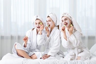 Photo of Happy women with facial masks and cucumber slices in bathrobes after spa procedures on bed indoors