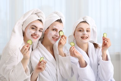 Photo of Happy women with facial masks and cucumber slices in bathrobes after spa procedures indoors