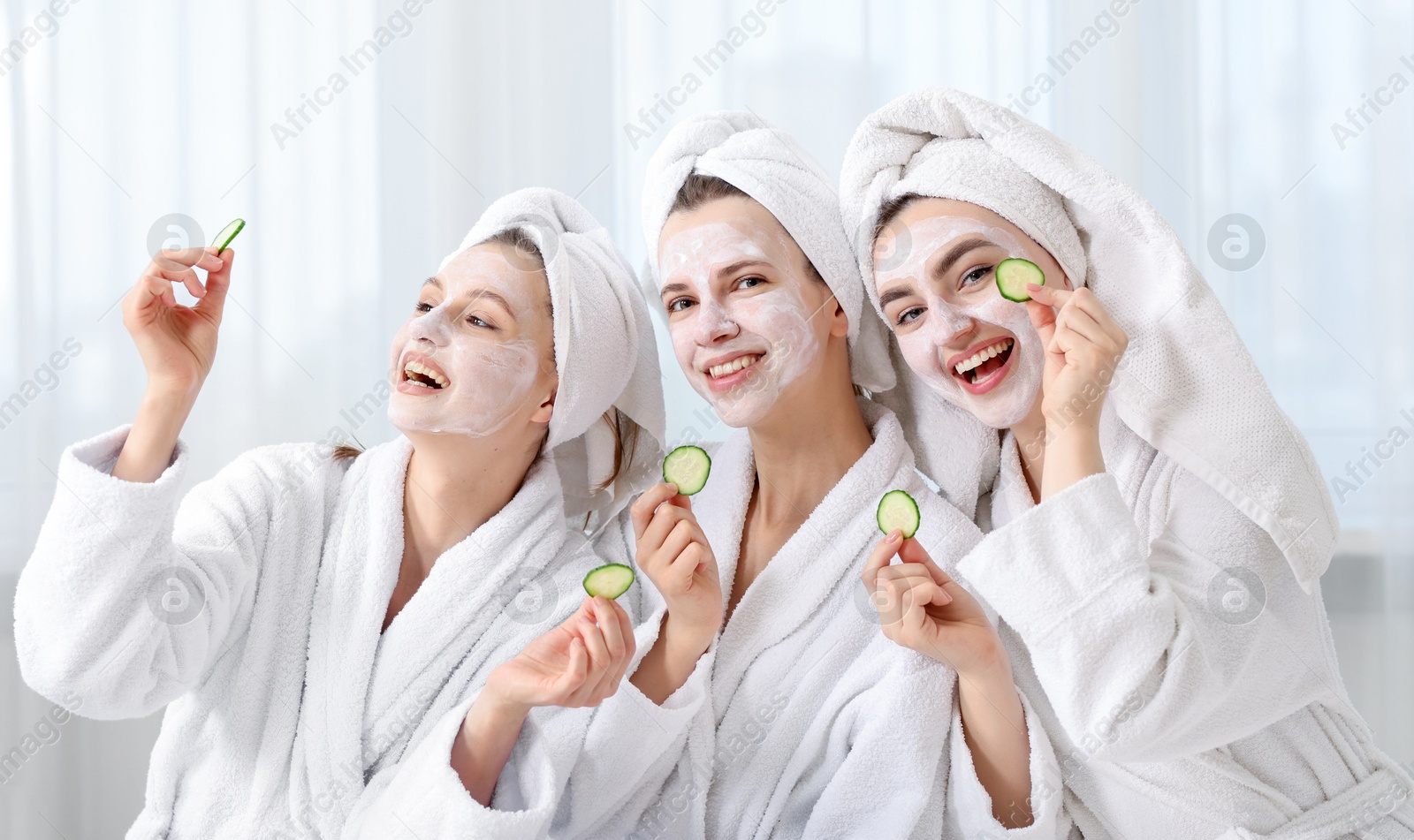 Photo of Happy women with facial masks and cucumber slices in bathrobes after spa procedures indoors