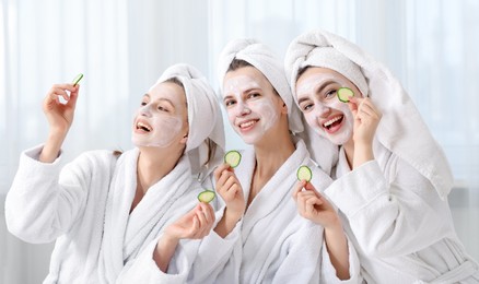 Photo of Happy women with facial masks and cucumber slices in bathrobes after spa procedures indoors