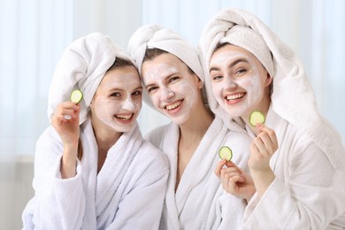 Photo of Happy women with facial masks and cucumber slices in bathrobes after spa procedures indoors