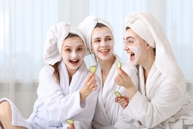 Photo of Happy women with facial masks and cucumber slices in bathrobes after spa procedures indoors
