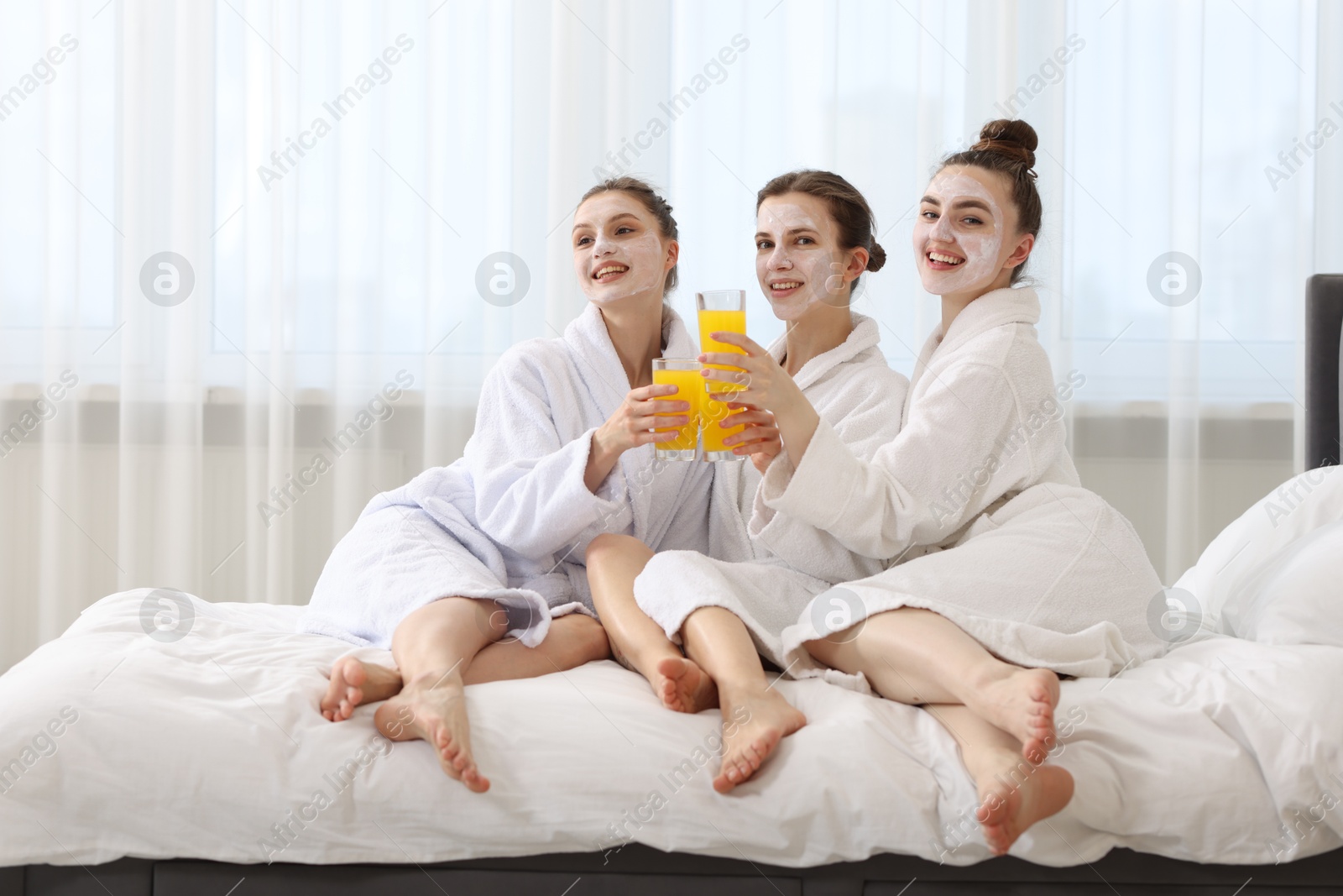 Photo of Happy women with facial masks and glasses of orange juice in bathrobes after spa procedures on bed indoors