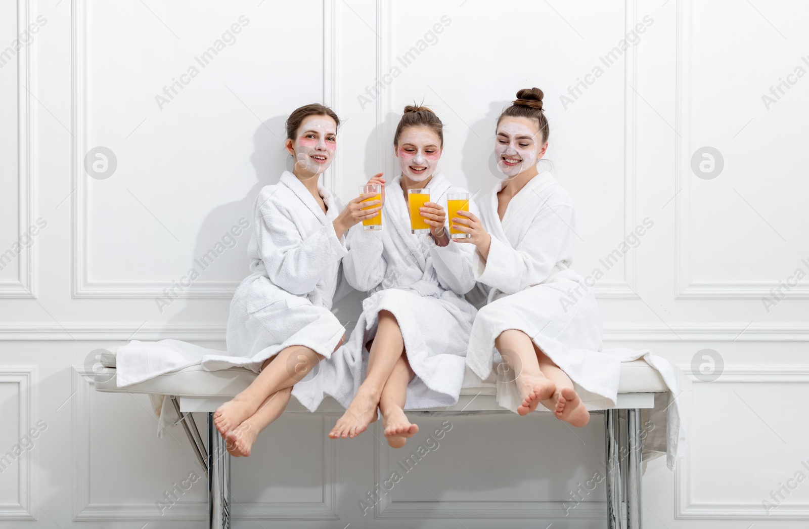 Photo of Happy women with facial masks and glasses of orange juice in bathrobes after spa procedures sitting on massage couch near white wall