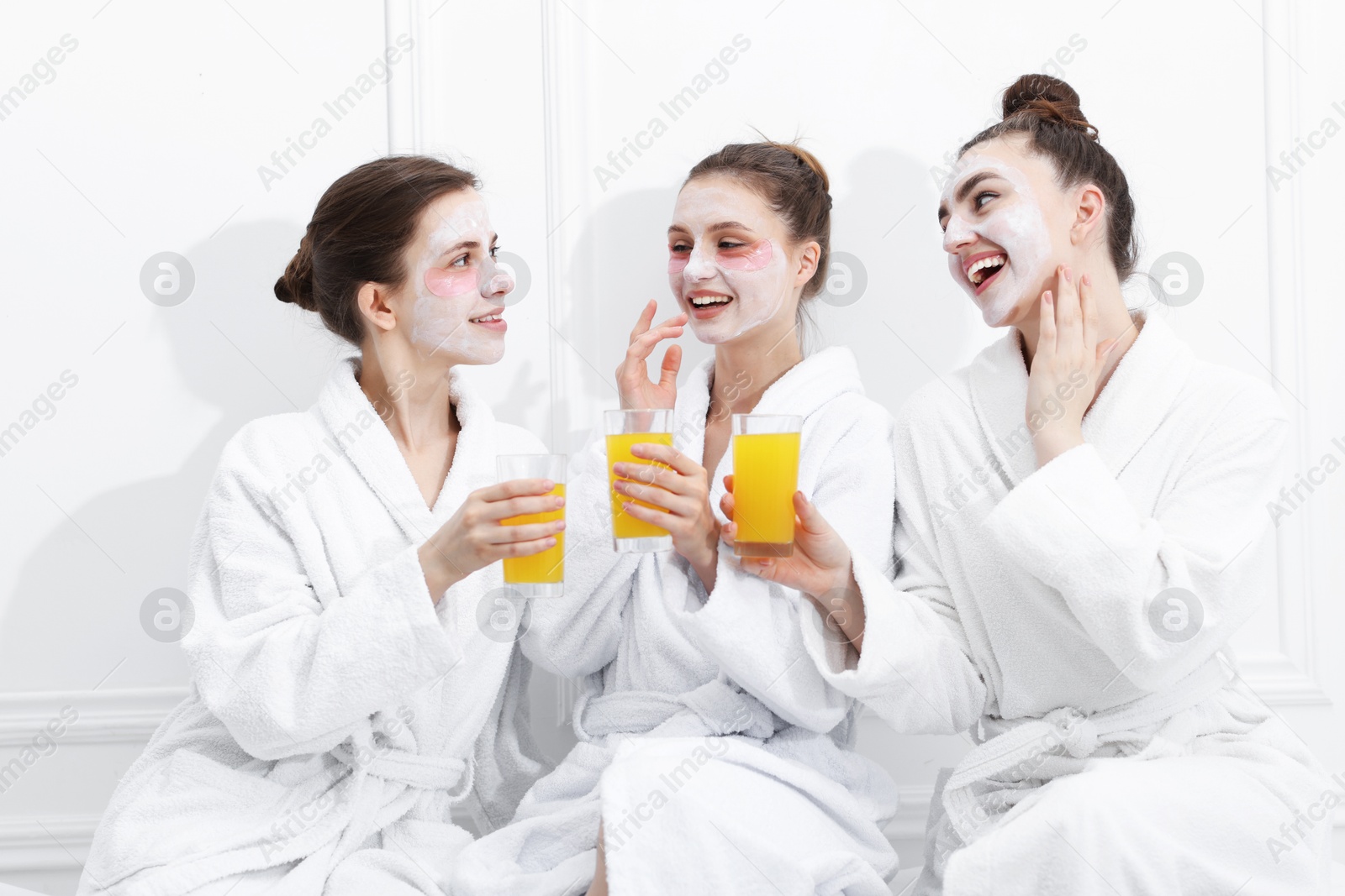 Photo of Happy women with facial masks and glasses of orange juice in bathrobes after spa procedures near white wall