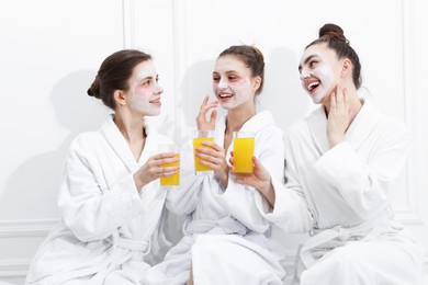 Photo of Happy women with facial masks and glasses of orange juice in bathrobes after spa procedures near white wall