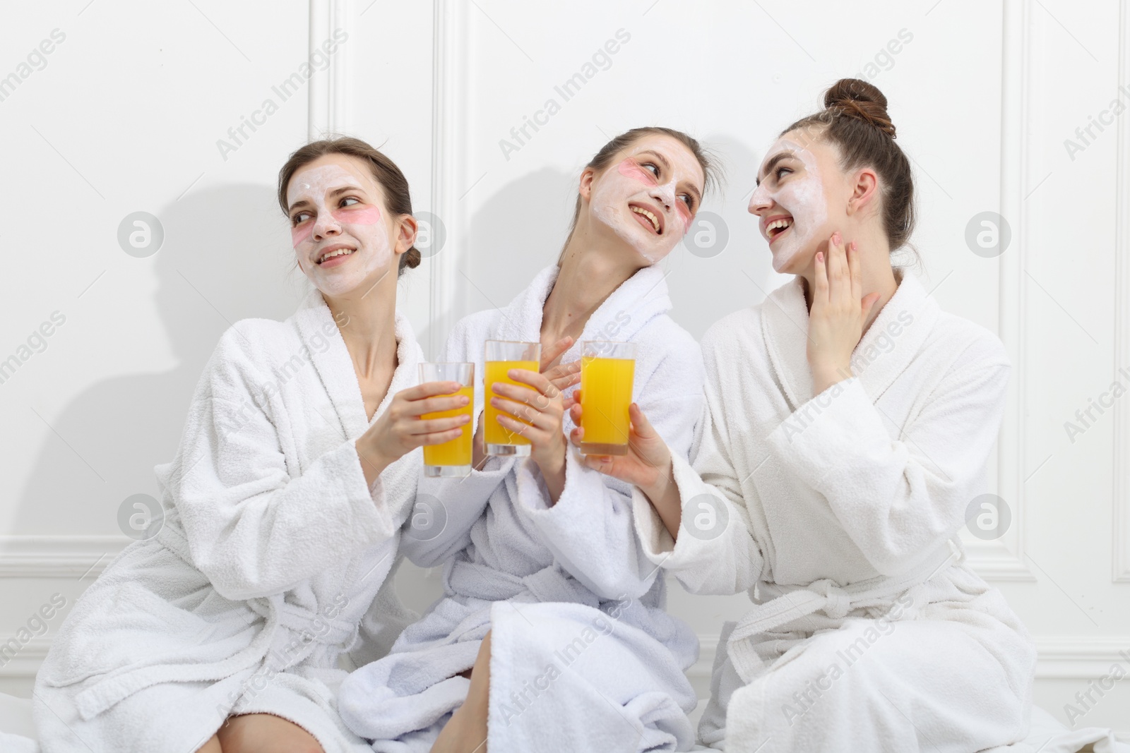 Photo of Happy women with facial masks and glasses of orange juice in bathrobes after spa procedures near white wall