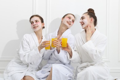 Photo of Happy women with facial masks and glasses of orange juice in bathrobes after spa procedures near white wall