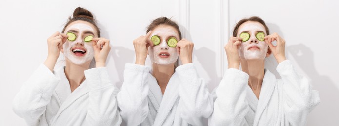 Photo of Happy women with facial masks and cucumber slices in bathrobes after spa procedures near white wall