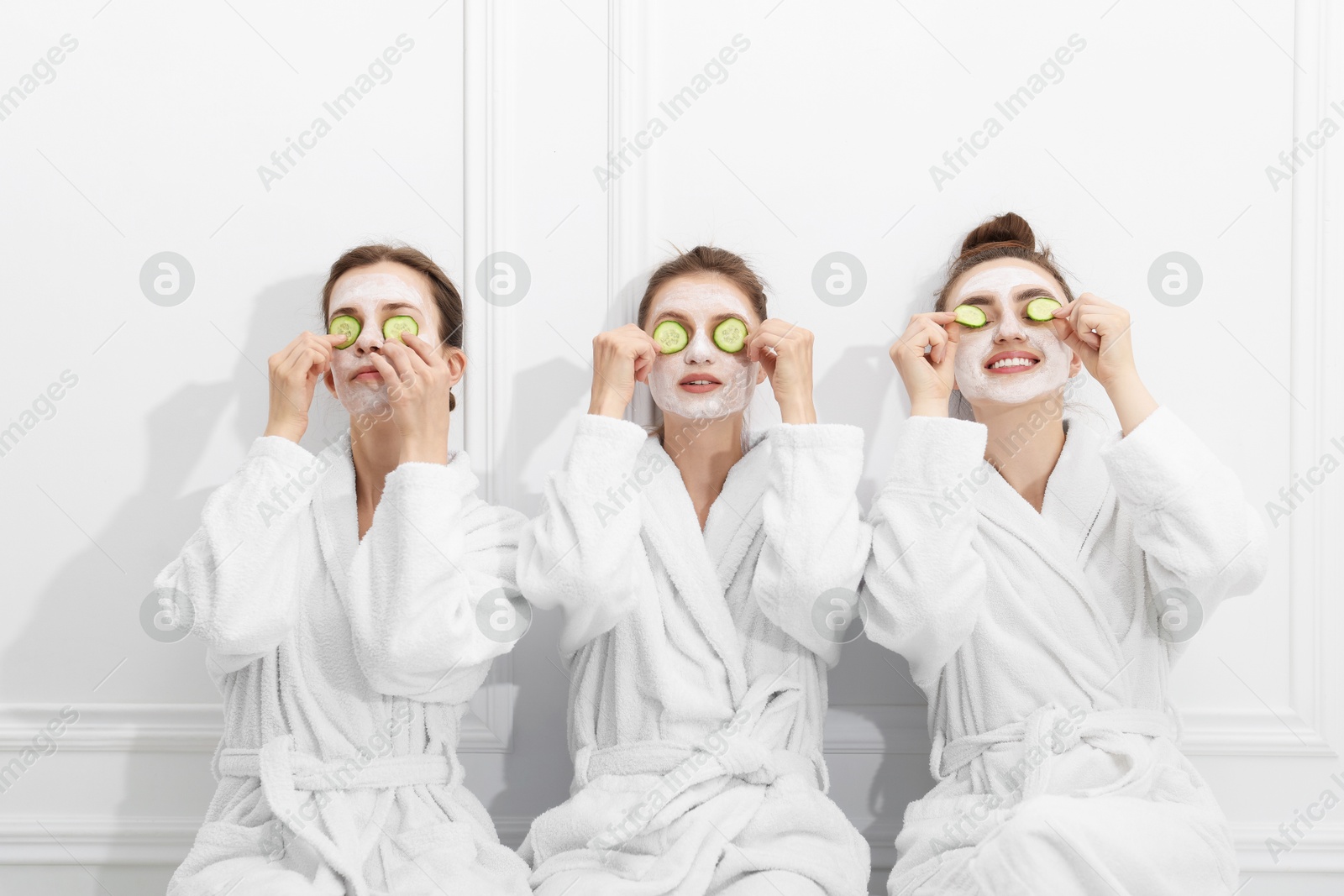 Photo of Happy women with facial masks and cucumber slices in bathrobes after spa procedures near white wall