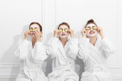Photo of Happy women with facial masks and cucumber slices in bathrobes after spa procedures near white wall