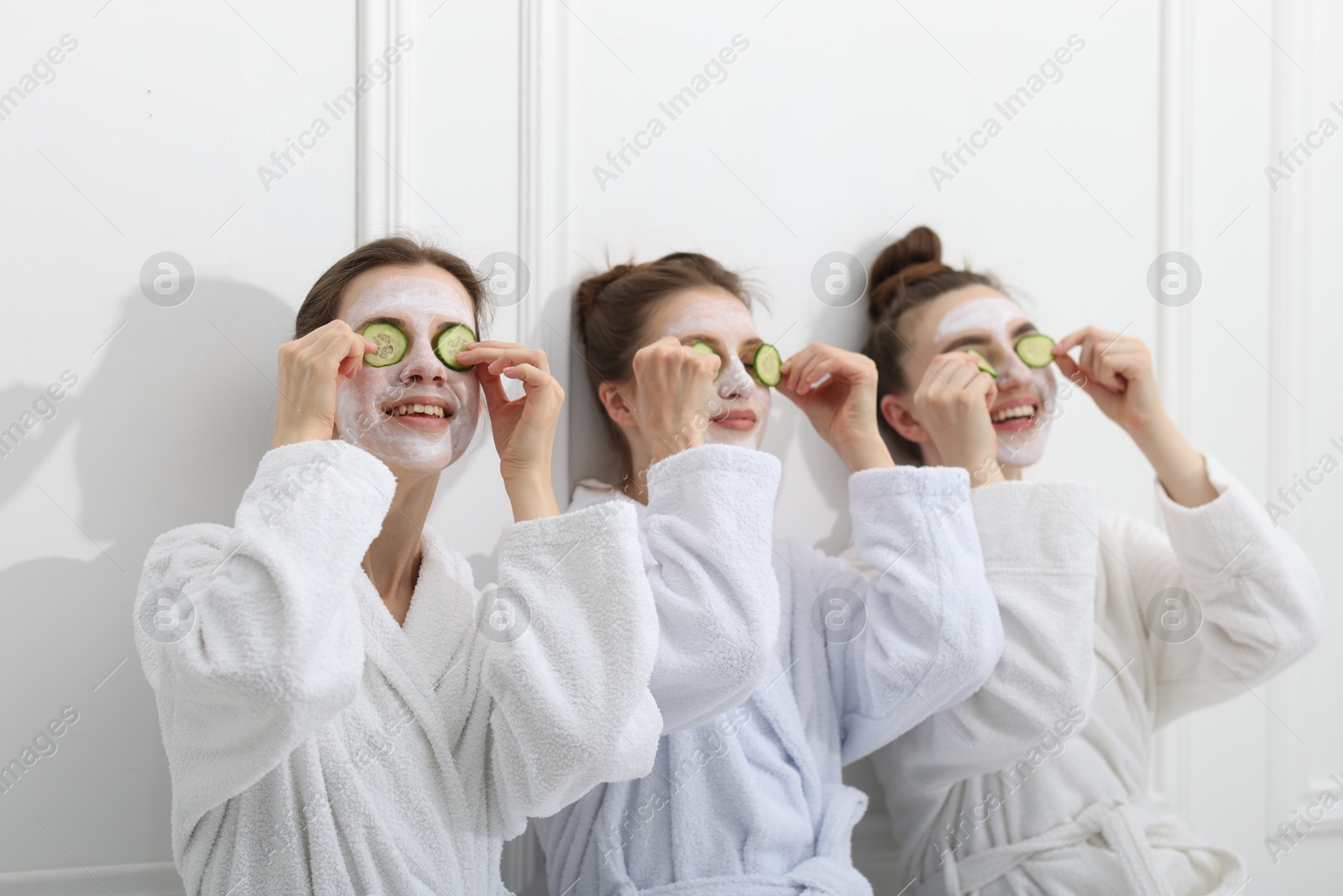 Photo of Happy women with facial masks and cucumber slices in bathrobes after spa procedures near white wall