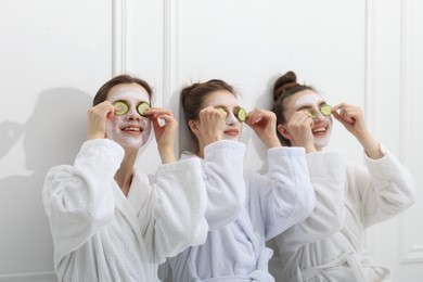 Photo of Happy women with facial masks and cucumber slices in bathrobes after spa procedures near white wall