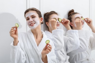 Photo of Happy women with facial masks and cucumber slices in bathrobes after spa procedures near white wall, selective focus